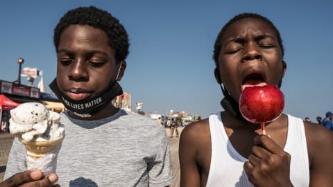 A street photograph on a boardwalk on a sunny day. Taking up the foreground are two young Black men side by side. The one on the left is looking down at the ice cream cone he is holding. The one on the right is in mid-bite of a red candy apple.