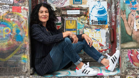 A full body portrait of artist Jannette Beckman. She is seated in the nook of a graffiti wall.