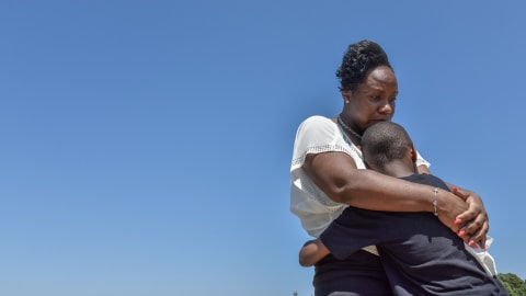 An African-American woman hugs her child. The background is a plain blue sky. She looks sad. We cannot see his face.