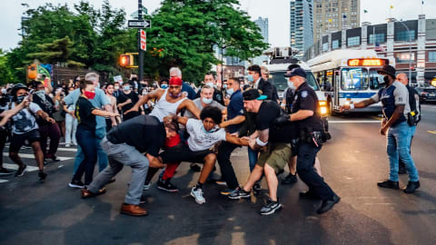 This photo taken of a Black Lives Matter protest is in the middle of a city street, with a confrontation between civilians and police officers. The main subject is an African-American man being tugged by his arm and leg on all sides.