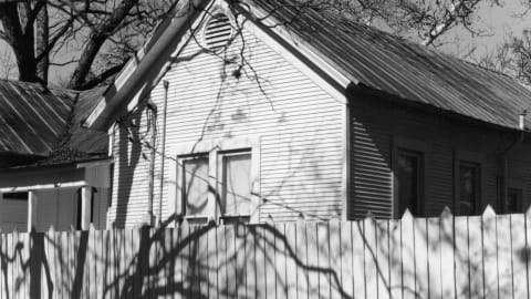 A black-and-white photograph of a fence and a house, with the shadow of tree branches falling over the fence.