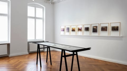 Color photograph of a gallery room with white walls and two windows on the left. On the right wall there is a rail displaying transparencies in frames. In the middle of the space there is a display table with images and text. 