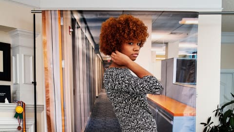 A young black woman with curly hair stands in a domestic living room front of a printed background representing a typical office space with cubicles.