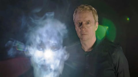 A man in a black shirt standing in front of an illuminated white smoke in the shape of a face
