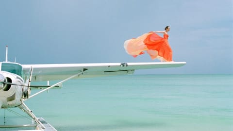 Alt Text: A woman in an orange dress standing on the edge of the wing of a sea plane. She is surrounded by turquoise ocean water and her dress is blowing dramatically in the wind.