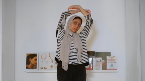 A woman stands on a white bed with her arms circled over her head. She is wearing a headscarf, striped shirt and black pants.