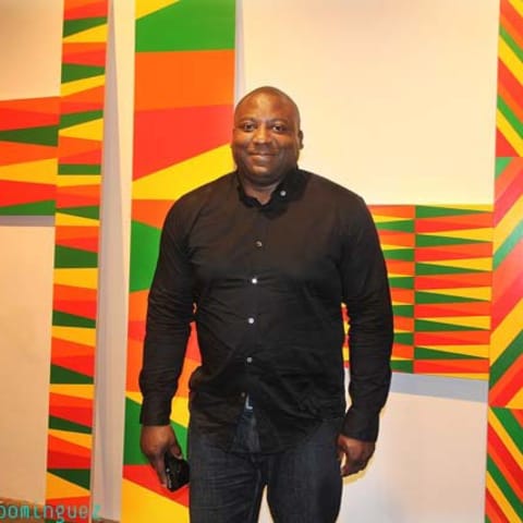 Rico Gatson in a black dress shirt standing in front of a colorfully patterned artwork