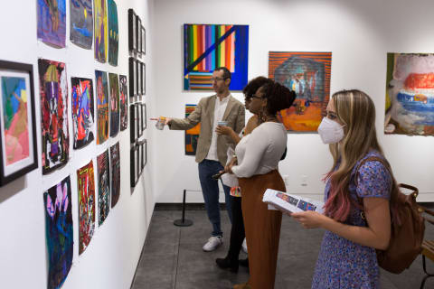 Three women and a man look at a wall on which is installed many small paintings.