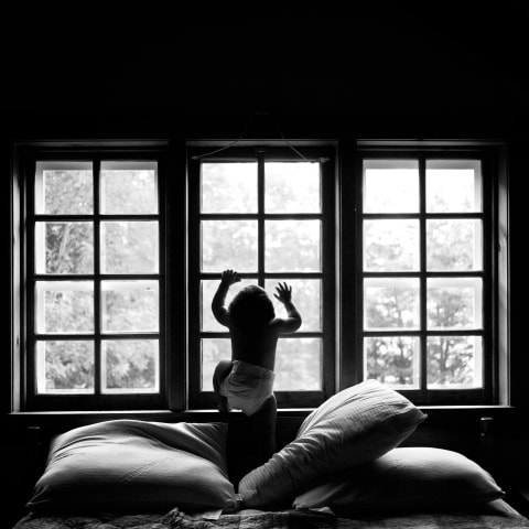  A black and white image of a window seat with large pillows underneath a three-panel window. There is a baby attempting to climb up the middle window and is silhouetted from the light coming through the window behind him.