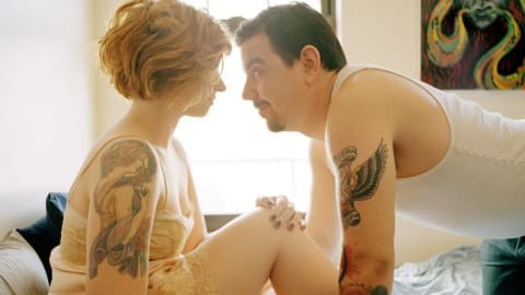 A man and a woman, both tattooed, share an intimate moment on a bed.
