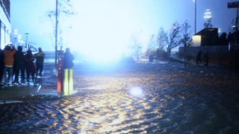 Flooded street at night with a bright light on the horizon. People are standing on the sidewalks.