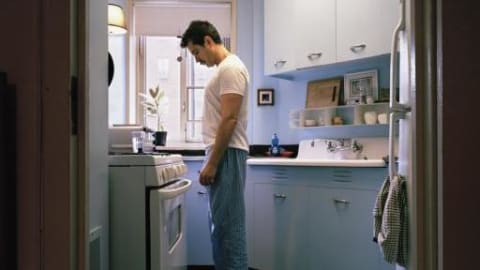 A pajama clad man is standing on a scale inside of a clean, apartment kitchen with his slippers on the ground behind him.