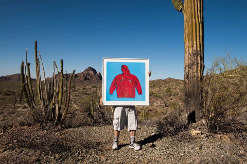 Photographer Tom Kiefer near his studio in in Ajo, Arizona, in the desert with a huge cactus next to him. He is holding up one of his framed photos of a red hooded sweater on a blue background. The letters on the sweater say MICKEY.
