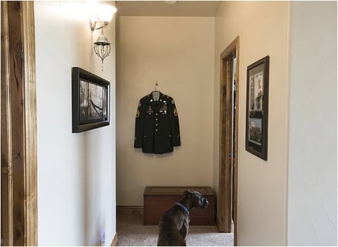 A dog sitting in the hall way in front of an army jacket.