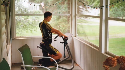 Photograph of a light-skinned man on exercise bike looking at a list of movements while inside an enclosed patio or sunroom.