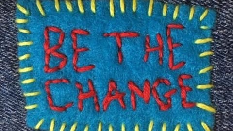 A blue square of fabric with yellow stitching around the borders. The words Be The Change, are stitched in red within the square