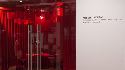 View of "The Red Room" installation through glass doors of gallery, with "The Red Room, an exhibition by BFA Interior Design" and the exhibition dates in vinyl lettering to wall on far right side of frame.
