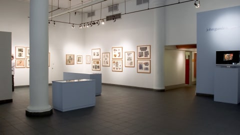 Lobby gallery view from inside Visual Arts Museum, showing multiple framed drawings on left side of room, and two vitrines in center of space, with exhibition text “John Gundlefinger” on wall to the right and a monitor playing a slideshow of artwork.
