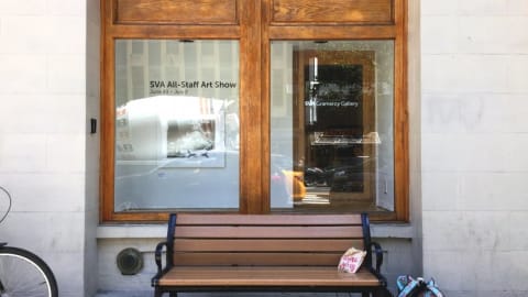 A bench in front of a window with a bike to the left and a bag to the right on the ground with a reflection of a cement mixer in the window.