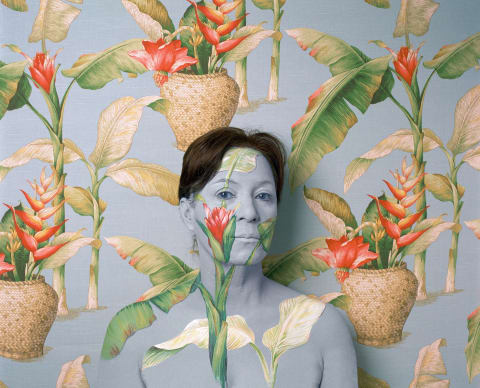A photograph of a woman from the chest up standing in front of an ornately-wallpapered wall. The wallpaper is pale blue, and there are green plants with orange flowers sticking out of woven pots. The woman is painted with the same pattern to blend into the wallpaper, with only her brown hair standing out.