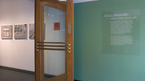 Photo from just inside main doors at Visual Arts Museum, showing the exhibition title and curator’s statement on wall to the left, then just inside the wood and glass entry doors are three paintings.