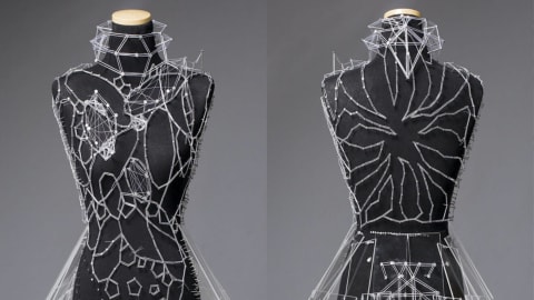A delicately designed dress, with white thread and clear plexiglass panels, on a black dress form.