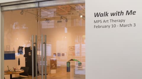 A view from outside of a shop's entrance with transparent glass doors and a write on a wall "Walk with Me-MPS Art therapy February 10-March 3"