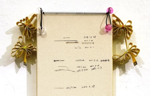Image is the top half of a sculpture made of a notebeook with tassels and ribbons on the sides.  Written on the notebook are different measurements.