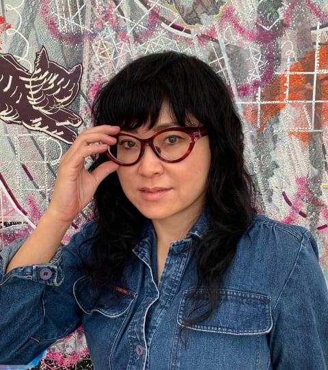 A photograph of artist, Chie Fueki, standing in front of one of her artworks. Chie is an Asian female with round glasses and a blue denim shirt. Chie is looking into the camera while holding up her right hand to her glasses.