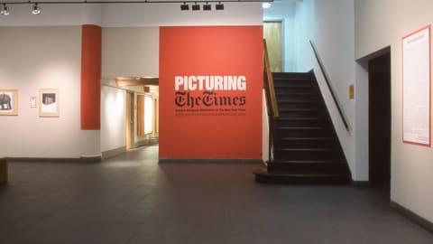 Lobby gallery at Visual Arts Museum showing two framed artworks on the left wall and in the center of the space is a red-painted wall with the exhibition title “Picturing The Times” next to a staircase.