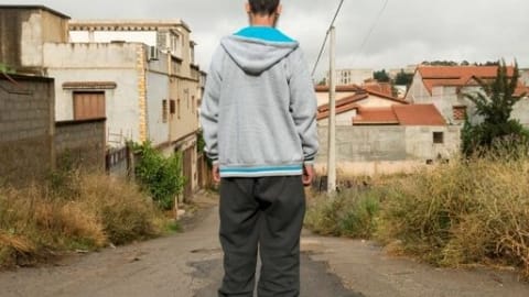 A man with baggy pants and sweatshirt looks down the street in a residential neighborhood of masonry buildings.