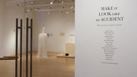 Photograph from outside SVA Flatiron Gallery looking in through glass doors, with exhibition title "Make It Look Like An Accident" displayed on the wall to the right.