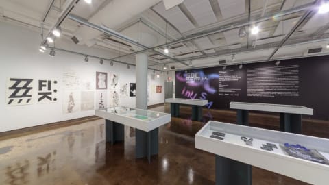 Photograph of first gallery room with four vitrines containing objects in the center of the room, a large black poster displaying exhibition information on the back right wall, and on the back left wall are many different poster designs.