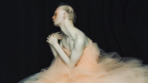 Photograph of a light-skinned person with very short blond hair dressed in a peach tulle dress, standing in profile against black fabric backdrop.