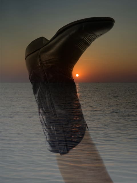 Black Boot: Multiple exposure on lower leg wearing black cowboy boot extending over sunset and seaside.