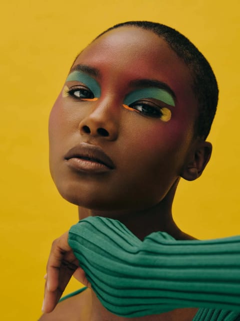 A fashion portrait of a black woman wearing colorful makeup and a green sweater.