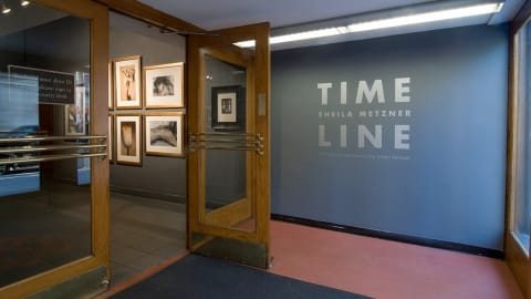 Photograph from just outside lobby gallery of Visual Arts Museum with exhibition text, "Time Line: Sheila Metzner" on the wall to the right, on the left inside gallery are several framed photographs.