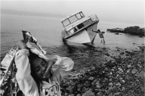 Black and white photograph of boat tipped slightly to its side in the shallow water off a pebbly beach. A boy is standing in the water leaning on the boat. In the foreground and out of focus a girl is laughing with one hand on her mouth and holding a silver blanket in the other.