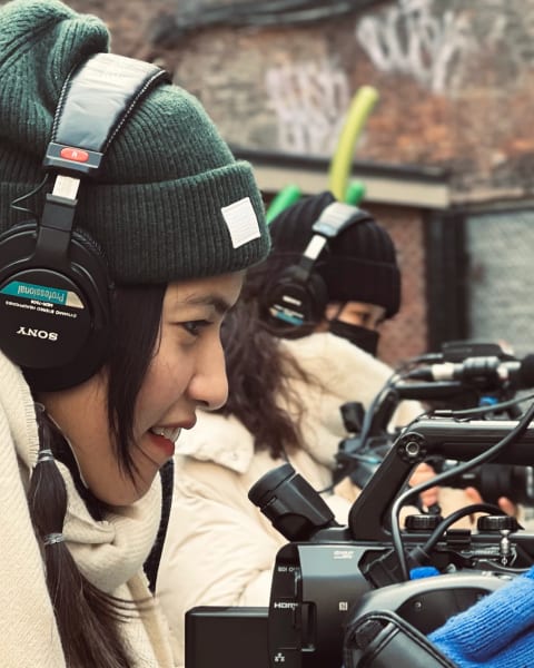 a woman films outside, wearing hat, gloves & headphones and in profile facing right. She looks into her camera. Another woman in a similar position is seen behind her.