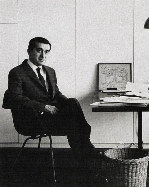 a black and white photo; a man in suit, sitting by a desk with a lamp looking at the camera. 