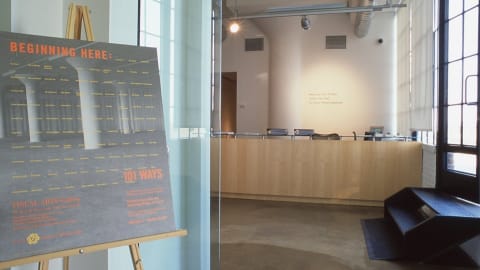 Photograph taken outside of Visual Arts Gallery, showing promotional sign with exhibition details to the left, and on the right is a view of the reception desk inside gallery.