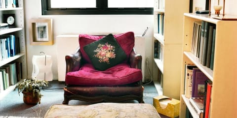 Photo of a room with a red chair that has a floral pillow placed on it.  The chair is surrounded by bookshelves on the right and left, a plant and a wall and bottom of a window in the background.