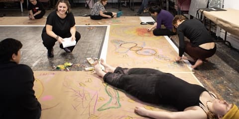 people drawing on a paper taped to the ground.  One person laying down and another crouched down and smiling holding paper.
