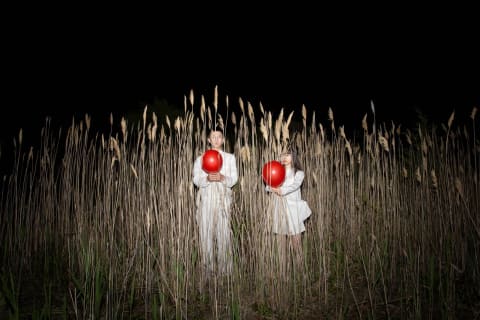 A man and woman wearing all white are each holding a red balloon, standing in tall reeds at night.