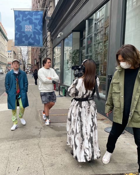 5 students walk towards the camera, 2 walk backwards holding cameras, filming another two. A blue SVA NYC banner flies in the background