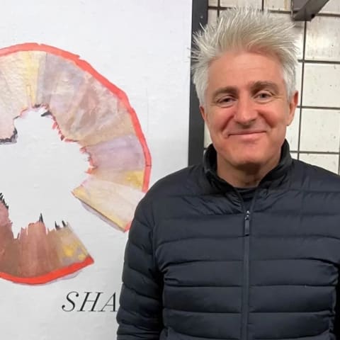 A man with gray hair, wearing a black jacket, smiling as he stands next to a poster in the subway. 