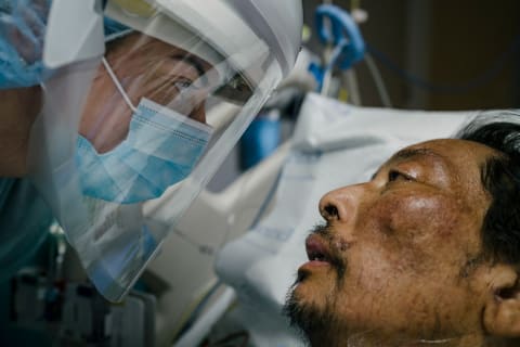 A female healthcare worker wearing personal protective equipment leans towards her patient’s face and makes eye contact with him, as he lies in bed, in an intensive care unit.