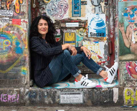 A full body portrait of artist Jannette Beckman. She is seated in the nook of a graffiti wall.