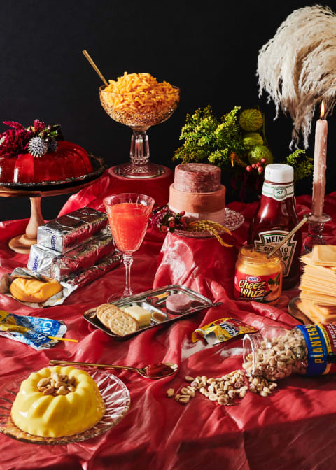 A photograph of a still life on a table with a red tablecloth and a black background. On it are various foods; peanuts spilling out of a bottle, cheez whiz, capri sun, Velveeta, kool-aid, ketchup, crackers, jello, stacked salami, and a glass bowl of mac and cheese
