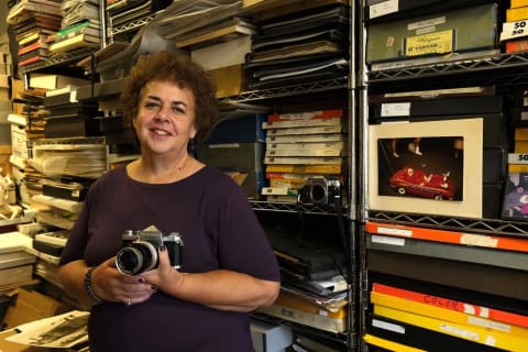 Portrait of Mary Engel taken in front of shelves of photo archives. She is holding a camera and smiling.